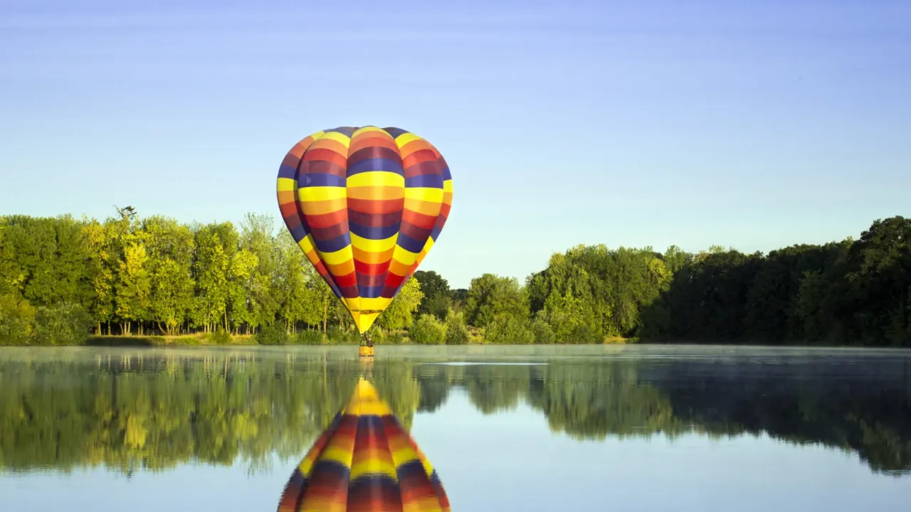 Montgolfière colorée survolant un lac paisible avec reflet dans l'eau au lever du soleil.