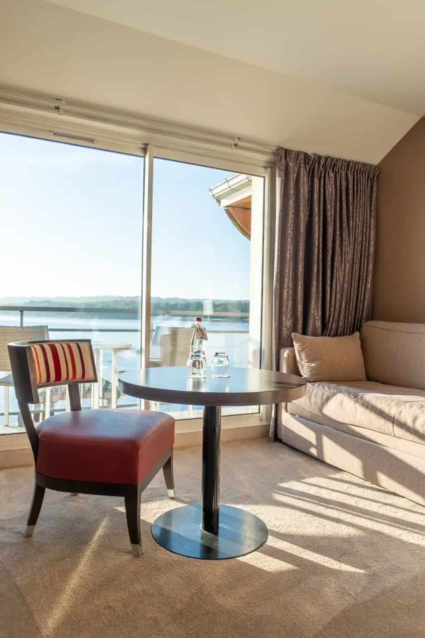 Coin salon lumineux d'une chambre avec table, chaise rouge et vue sur l'extérieur.